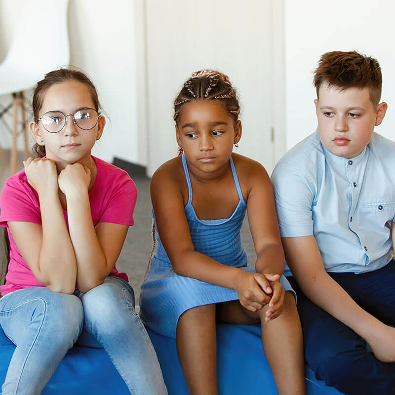 Three sad, serious teenagers are sitting. Caucasian, African American, boy and girl with glasses.